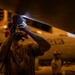 A maintenance technician is inspecting the underside of a military aircraft using a handheld flashlight in a low-light environment, likely inside an aircraft carrier hangar bay. The aircraft bears the number '213' on its fuselage. A second crew member wearing a helmet is visible in the background.