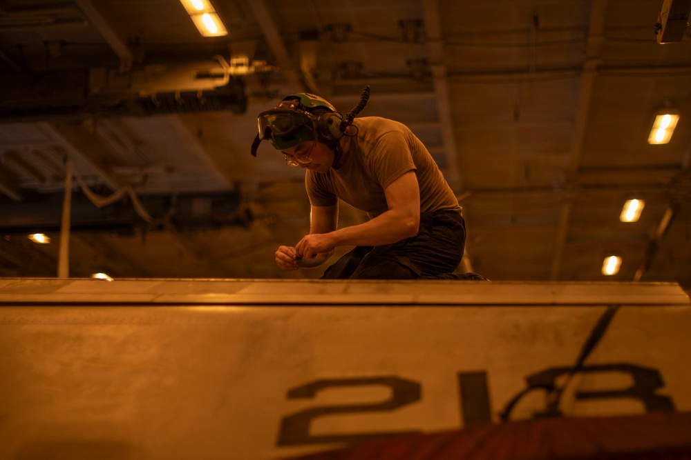 A military or naval technician is crouched on top of a large piece of equipment or vehicle, marked with the number '218', working with their hands in what appears to be a hangar bay or below-deck space. The environment is dimly lit with warm amber/orange overhead lighting, consistent with a naval vessel's interior. The worker is wearing protective goggles pushed up on their head and a communications headset.