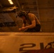 A military or naval technician is crouched on top of a large piece of equipment or vehicle, marked with the number '218', working with their hands in what appears to be a hangar bay or below-deck space. The environment is dimly lit with warm amber/orange overhead lighting, consistent with a naval vessel's interior. The worker is wearing protective goggles pushed up on their head and a communications headset.