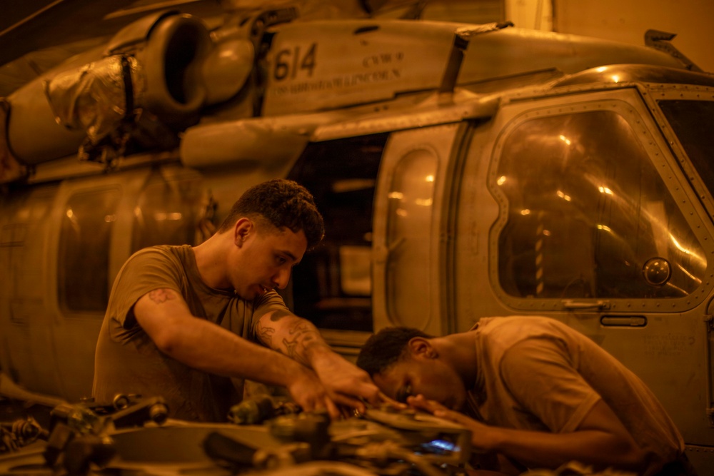 Two military personnel in tan/olive uniforms are performing maintenance work on a military helicopter in what appears to be a hangar or ship deck bay. The scene is illuminated by warm amber/orange artificial lighting, creating a low-light working environment. The helicopter bears the number '614' and appears to be a Sikorsky SH-60 Seahawk or similar naval helicopter.