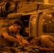 Two military personnel in tan/olive uniforms are performing maintenance work on a military helicopter in what appears to be a hangar or ship deck bay. The scene is illuminated by warm amber/orange artificial lighting, creating a low-light working environment. The helicopter bears the number '614' and appears to be a Sikorsky SH-60 Seahawk or similar naval helicopter.