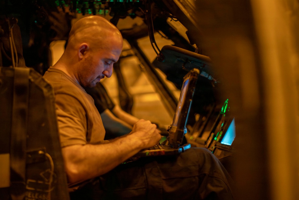A bald man wearing a tan/olive military-style t-shirt is seated inside what appears to be a military aircraft or vehicle, working intently on a laptop or keyboard device. The environment is dimly lit with warm amber and green indicator lights visible in the background. The setting suggests an operational or technical work environment, likely airborne or in a tactical vehicle.