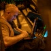 A bald man wearing a tan/olive military-style t-shirt is seated inside what appears to be a military aircraft or vehicle, working intently on a laptop or keyboard device. The environment is dimly lit with warm amber and green indicator lights visible in the background. The setting suggests an operational or technical work environment, likely airborne or in a tactical vehicle.