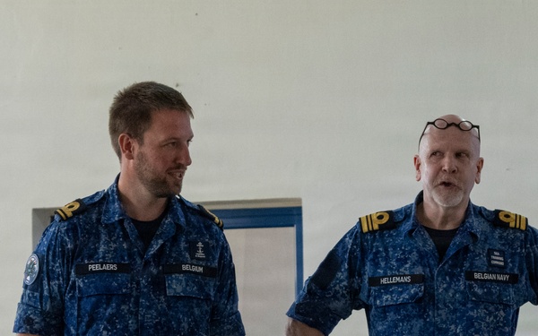 Four military personnel are engaged in conversation in what appears to be a meeting or dining facility. Two individuals wearing US Navy digital camouflage uniforms (NWU Type III) are speaking with two members of the Belgian Navy in blue digital camouflage uniforms. Name tapes visible on the Belgian Navy personnel read 'PEELAERS' and 'HELLEMANS', both with 'BELGIUM' or 'BELGIAN NAVY' identifiers.