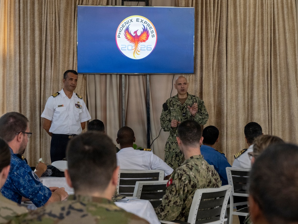 A military briefing or conference is taking place under the 'Phoenix Express 2026' exercise banner, with two uniformed officers standing at the front of the room addressing an audience of seated military personnel. The event appears to be a multinational naval exercise planning or orientation session, with participants wearing various national military uniforms. One officer in U.S. Navy digital camouflage (NWU) is speaking while another in white naval dress uniform stands nearby.