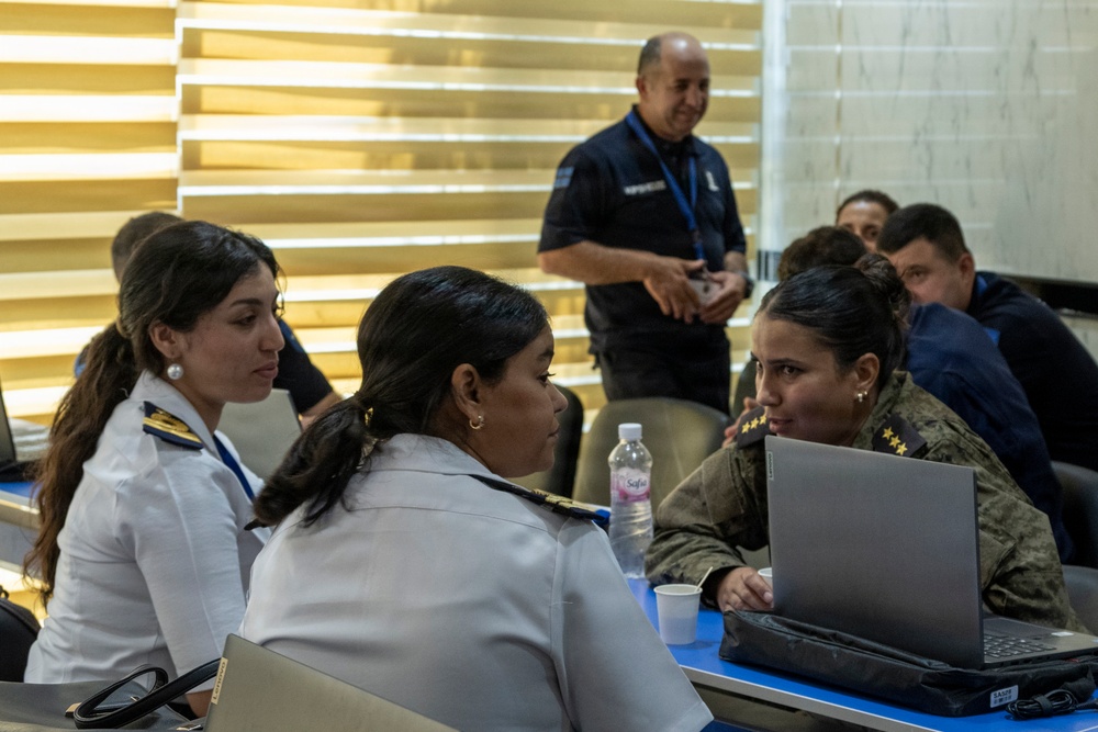 A group of uniformed military and law enforcement personnel are gathered around a table in what appears to be a conference or briefing room. Three women in the foreground are engaged in conversation, with one using a Lenovo laptop. Additional personnel are visible in the background, including a man in a dark uniform with a blue lanyard standing and smiling.
