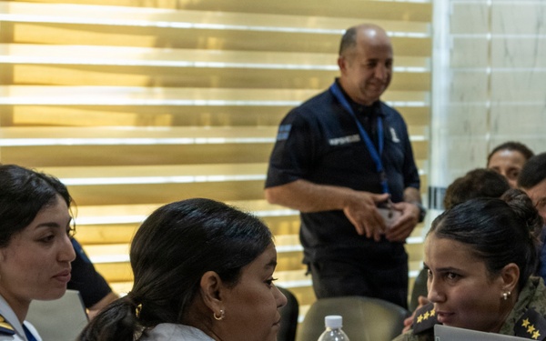 A group of uniformed military and law enforcement personnel are gathered around a table in what appears to be a conference or briefing room. Three women in the foreground are engaged in conversation, with one using a Lenovo laptop. Additional personnel are visible in the background, including a man in a dark uniform with a blue lanyard standing and smiling.