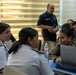 A group of uniformed military and law enforcement personnel are gathered around a table in what appears to be a conference or briefing room. Three women in the foreground are engaged in conversation, with one using a Lenovo laptop. Additional personnel are visible in the background, including a man in a dark uniform with a blue lanyard standing and smiling.