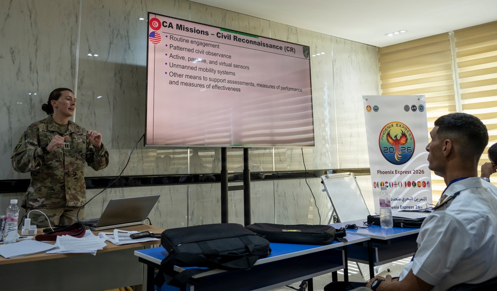 A U.S. military service member in camouflage uniform is delivering a briefing on 'CA Missions – Civil Reconnaissance (CR)' to an audience that includes at least one foreign military officer. The presentation is part of 'Phoenix Express 2026,' a multinational naval exercise, as indicated by the banner visible in the background. The setting appears to be a formal conference or classroom environment.