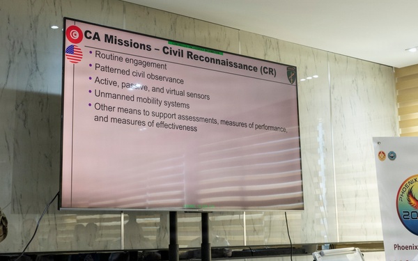 A U.S. military service member in camouflage uniform is delivering a briefing on 'CA Missions – Civil Reconnaissance (CR)' to an audience that includes at least one foreign military officer. The presentation is part of 'Phoenix Express 2026,' a multinational naval exercise, as indicated by the banner visible in the background. The setting appears to be a formal conference or classroom environment.