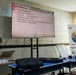 A U.S. military service member in camouflage uniform is delivering a briefing on 'CA Missions – Civil Reconnaissance (CR)' to an audience that includes at least one foreign military officer. The presentation is part of 'Phoenix Express 2026,' a multinational naval exercise, as indicated by the banner visible in the background. The setting appears to be a formal conference or classroom environment.
