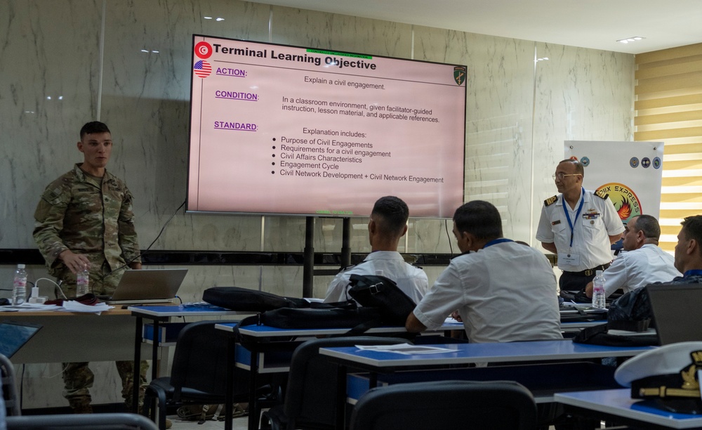 A military training or educational session is taking place in a classroom setting, with a U.S. Army soldier in camouflage uniform presenting a 'Terminal Learning Objective' slide about civil engagements. The audience consists of several military personnel in white naval/dress uniforms, and a senior officer in white uniform stands to the right near a banner. The presentation includes flags of Tunisia and the United States, suggesting a multinational military cooperation exercise.