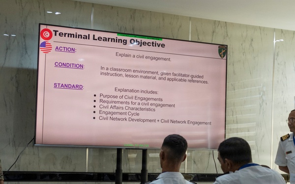 A military training or educational session is taking place in a classroom setting, with a U.S. Army soldier in camouflage uniform presenting a 'Terminal Learning Objective' slide about civil engagements. The audience consists of several military personnel in white naval/dress uniforms, and a senior officer in white uniform stands to the right near a banner. The presentation includes flags of Tunisia and the United States, suggesting a multinational military cooperation exercise.