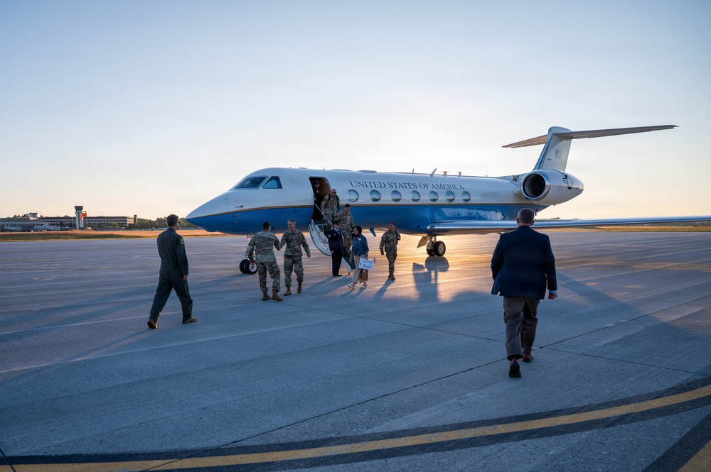 A United States of America government aircraft, likely a Gulfstream C-37, is parked on a tarmac at what appears to be a military airfield during golden hour. Military personnel in Army Combat Uniforms (ACU) are deplaning and being greeted by individuals on the tarmac, including a civilian in a suit and a person in a flight suit. The scene suggests an official arrival of senior military or government officials.