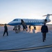 A United States of America government aircraft, likely a Gulfstream C-37, is parked on a tarmac at what appears to be a military airfield during golden hour. Military personnel in Army Combat Uniforms (ACU) are deplaning and being greeted by individuals on the tarmac, including a civilian in a suit and a person in a flight suit. The scene suggests an official arrival of senior military or government officials.