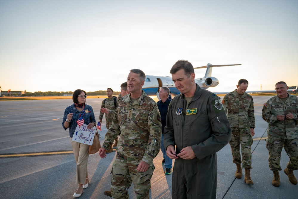 A group of U.S. Air Force military personnel are gathered on an airfield tarmac, apparently welcoming someone returning from a deployment or official trip. Two senior officers walk in the foreground, with a civilian woman holding a 'Welcome Home' sign visible in the background. A military transport aircraft (appears to be a C-21 or similar executive jet) is parked on the ramp behind the group.