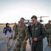 A group of U.S. Air Force military personnel are gathered on an airfield tarmac, apparently welcoming someone returning from a deployment or official trip. Two senior officers walk in the foreground, with a civilian woman holding a 'Welcome Home' sign visible in the background. A military transport aircraft (appears to be a C-21 or similar executive jet) is parked on the ramp behind the group.