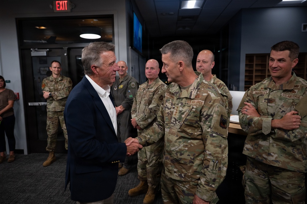 A civilian in a navy blazer is shaking hands with a U.S. Air Force officer in OCP (Operational Camouflage Pattern) uniform inside what appears to be a military facility or briefing room. Several other U.S. Air Force personnel in uniform are gathered in the background observing the exchange. The setting suggests an official visit or recognition ceremony.