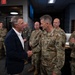 A civilian in a navy blazer is shaking hands with a U.S. Air Force officer in OCP (Operational Camouflage Pattern) uniform inside what appears to be a military facility or briefing room. Several other U.S. Air Force personnel in uniform are gathered in the background observing the exchange. The setting suggests an official visit or recognition ceremony.