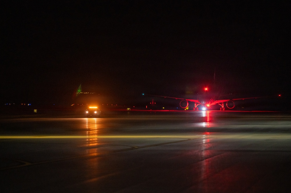A nighttime scene on an airport tarmac showing at least one large aircraft illuminated by red navigation/anti-collision lights, with a ground support vehicle nearby displaying amber/orange lights. The wet tarmac surface reflects the various colored lights, creating visible light streaks. A green navigation light is visible in the background, likely on another aircraft or structure.