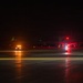 A nighttime scene on an airport tarmac showing at least one large aircraft illuminated by red navigation/anti-collision lights, with a ground support vehicle nearby displaying amber/orange lights. The wet tarmac surface reflects the various colored lights, creating visible light streaks. A green navigation light is visible in the background, likely on another aircraft or structure.