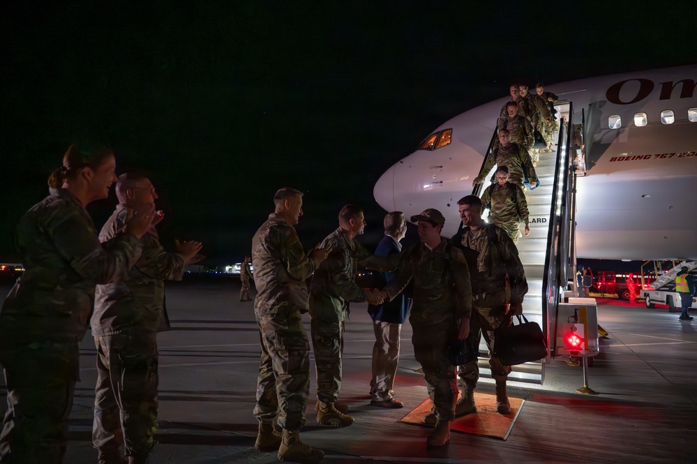 Military personnel are deplaning from a Boeing 767-200 series commercial charter aircraft at night on a tarmac. Returning service members in camouflage uniforms are being greeted by what appear to be senior officers and other personnel on the ground. The scene is consistent with a military unit homecoming or redeployment arrival ceremony.