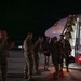 Military personnel are deplaning from a Boeing 767-200 series commercial charter aircraft at night on a tarmac. Returning service members in camouflage uniforms are being greeted by what appear to be senior officers and other personnel on the ground. The scene is consistent with a military unit homecoming or redeployment arrival ceremony.