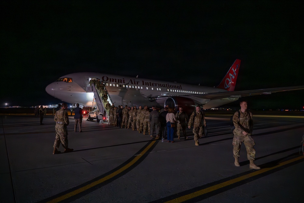 A nighttime scene on an airport tarmac showing military personnel deplaning from an Omni Air International commercial aircraft. A large group of uniformed service members in camouflage uniforms are gathered near the aircraft stairs and walking across the tarmac. The scene appears to depict a military deployment or redeployment operation.