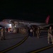 A nighttime scene on an airport tarmac showing military personnel deplaning from an Omni Air International commercial aircraft. A large group of uniformed service members in camouflage uniforms are gathered near the aircraft stairs and walking across the tarmac. The scene appears to depict a military deployment or redeployment operation.