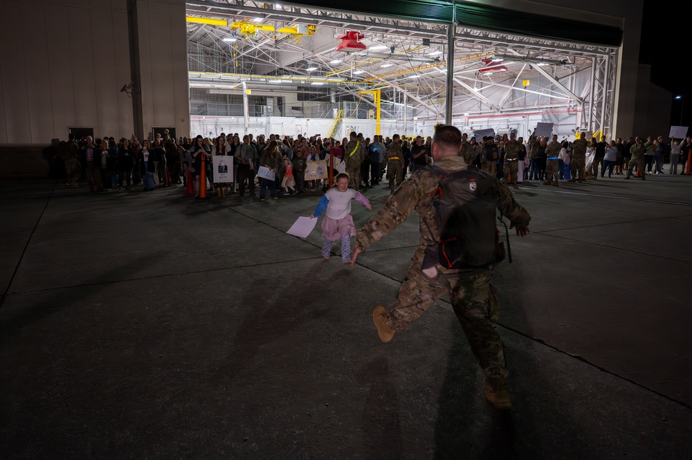 A military homecoming ceremony is taking place on an airfield tarmac at night, in front of a large illuminated hangar. A young child in pajamas is running toward a uniformed soldier who is rushing forward with arms outstretched to greet her. A large crowd of families, civilians, and military personnel holding welcome signs lines the background.