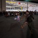 A military homecoming ceremony is taking place on an airfield tarmac at night, in front of a large illuminated hangar. A young child in pajamas is running toward a uniformed soldier who is rushing forward with arms outstretched to greet her. A large crowd of families, civilians, and military personnel holding welcome signs lines the background.