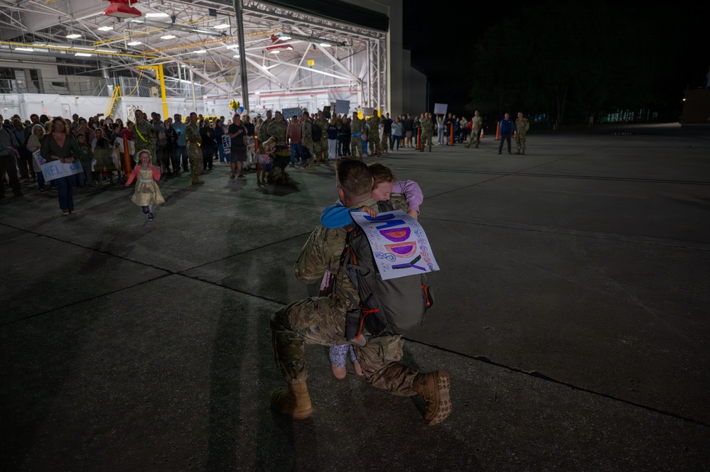 A military service member in camouflage uniform kneels on one knee on a tarmac at night, embracing a child who is holding a handmade welcome home sign. The reunion is taking place outside a large illuminated hangar where a crowd of families and other military personnel are gathered. This appears to be a military homecoming ceremony.