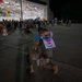 A military service member in camouflage uniform kneels on one knee on a tarmac at night, embracing a child who is holding a handmade welcome home sign. The reunion is taking place outside a large illuminated hangar where a crowd of families and other military personnel are gathered. This appears to be a military homecoming ceremony.