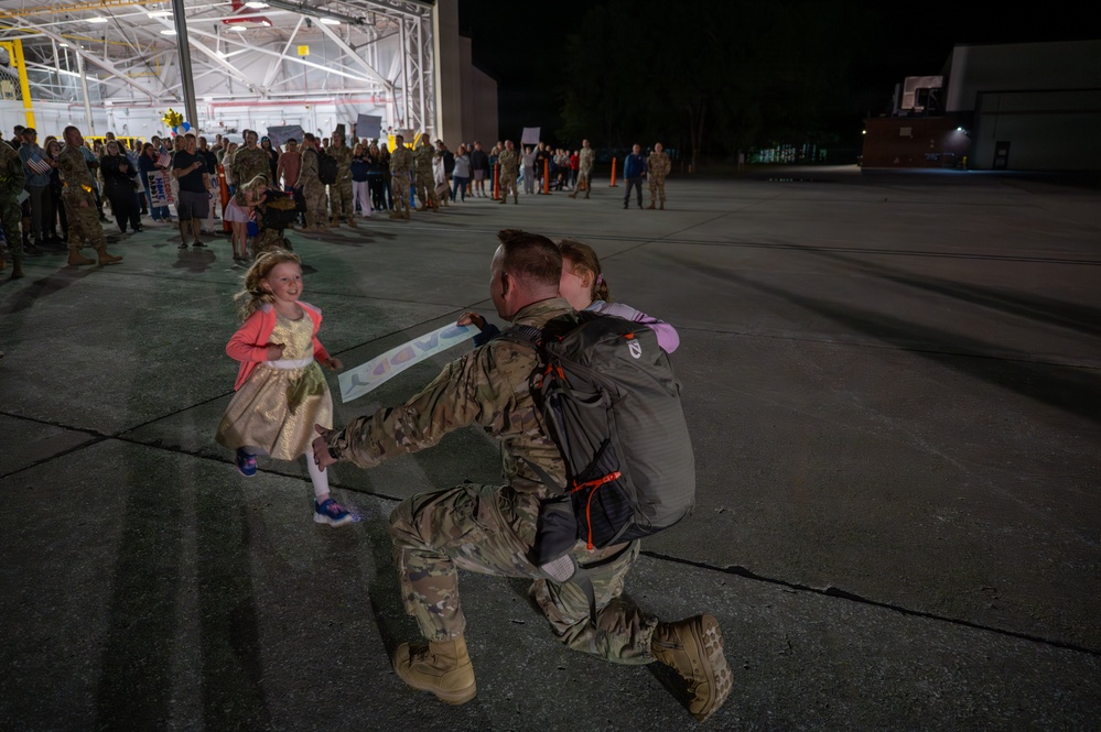 A military service member in camouflage uniform and full pack kneels on a tarmac to embrace a child while another young girl runs toward them smiling. The scene appears to be a military homecoming ceremony taking place at night outside a large hangar, with a crowd of uniformed personnel and civilians gathered in the background. The emotional reunion captures a soldier returning from deployment being greeted by children.