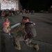 A military service member in camouflage uniform and full pack kneels on a tarmac to embrace a child while another young girl runs toward them smiling. The scene appears to be a military homecoming ceremony taking place at night outside a large hangar, with a crowd of uniformed personnel and civilians gathered in the background. The emotional reunion captures a soldier returning from deployment being greeted by children.