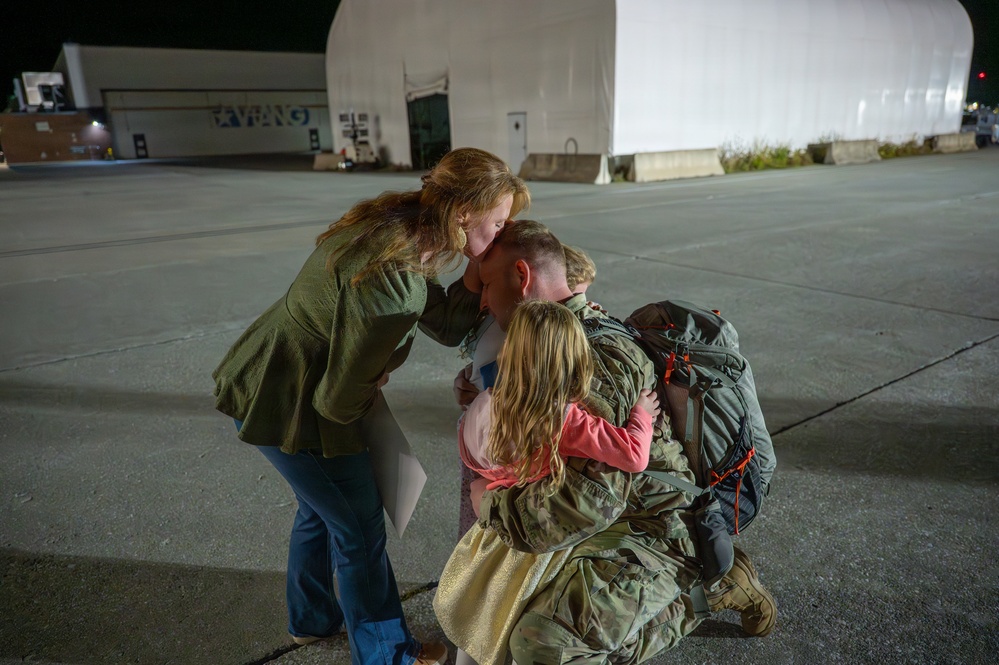 A military service member in camouflage uniform and tactical backpack kneels on a tarmac at night, being embraced by what appears to be a family — an adult woman and two young children. The setting appears to be a military air base or National Guard facility, indicated by the partial signage reading 'KYNG' on a building in the background. The scene strongly suggests a homecoming reunion.