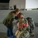 A military service member in camouflage uniform and tactical backpack kneels on a tarmac at night, being embraced by what appears to be a family — an adult woman and two young children. The setting appears to be a military air base or National Guard facility, indicated by the partial signage reading 'KYNG' on a building in the background. The scene strongly suggests a homecoming reunion.