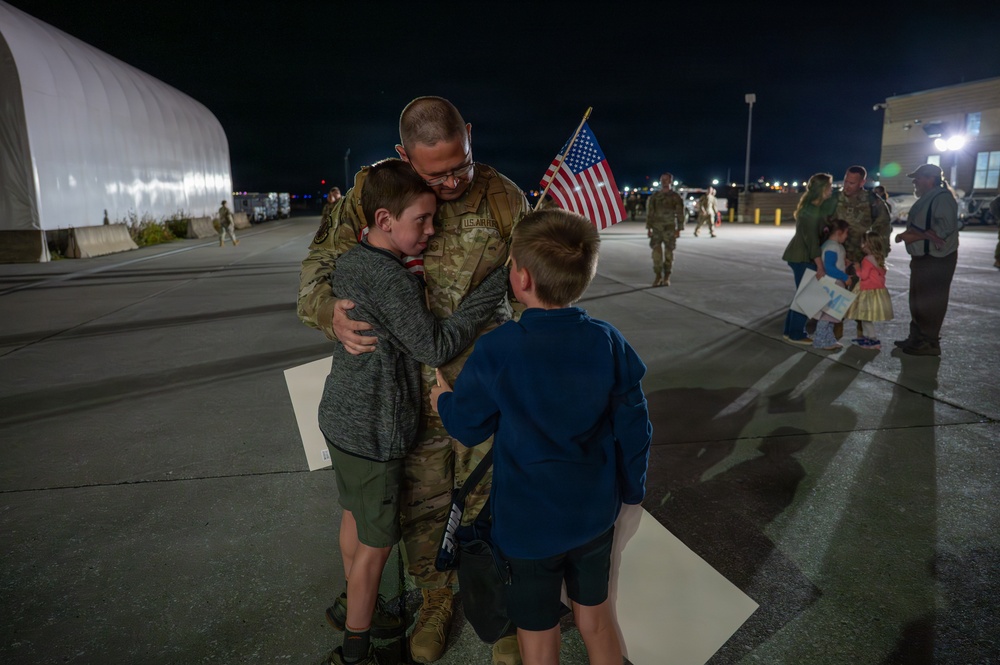 A U.S. military service member in camouflage uniform embraces two young boys on a tarmac at night, in what appears to be a homecoming or deployment farewell event. An American flag is visible being held by one of the children. In the background, other military personnel and civilians are gathered in similar reunions.