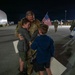 A U.S. military service member in camouflage uniform embraces two young boys on a tarmac at night, in what appears to be a homecoming or deployment farewell event. An American flag is visible being held by one of the children. In the background, other military personnel and civilians are gathered in similar reunions.