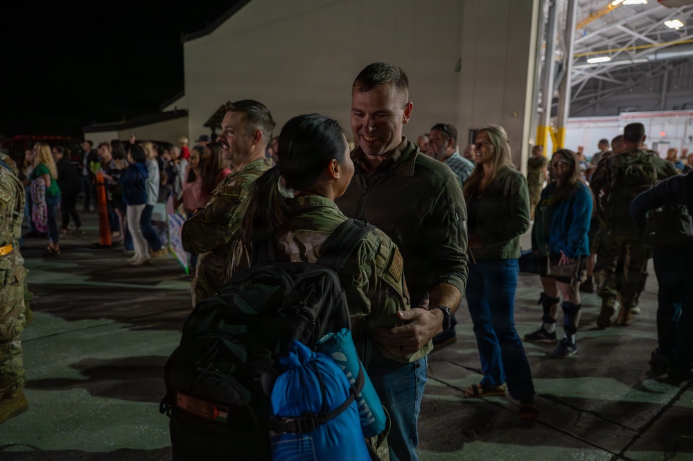 A military homecoming or deployment ceremony taking place on an airfield tarmac at night, near a large hangar. Uniformed service members are reuniting with civilians, with one central couple embracing in the foreground. The scene depicts an emotional gathering with numerous military personnel and their families or loved ones.