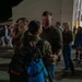 A military homecoming or deployment ceremony taking place on an airfield tarmac at night, near a large hangar. Uniformed service members are reuniting with civilians, with one central couple embracing in the foreground. The scene depicts an emotional gathering with numerous military personnel and their families or loved ones.