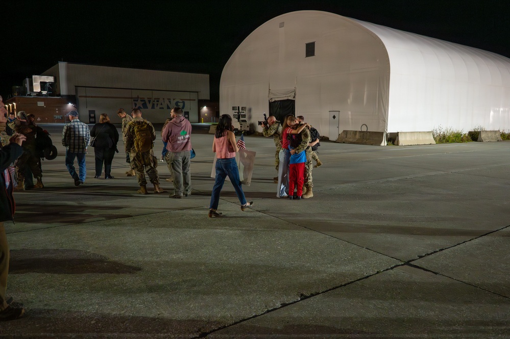 A nighttime military homecoming scene on an airfield tarmac, with service members in camouflage uniforms reuniting with family members. A central emotional moment is captured as a soldier embraces what appears to be family members while a photographer documents the reunion. A large white inflatable or fabric hangar structure is visible in the background alongside a brick building with partial signage.