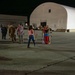A nighttime military homecoming scene on an airfield tarmac, with service members in camouflage uniforms reuniting with family members. A central emotional moment is captured as a soldier embraces what appears to be family members while a photographer documents the reunion. A large white inflatable or fabric hangar structure is visible in the background alongside a brick building with partial signage.