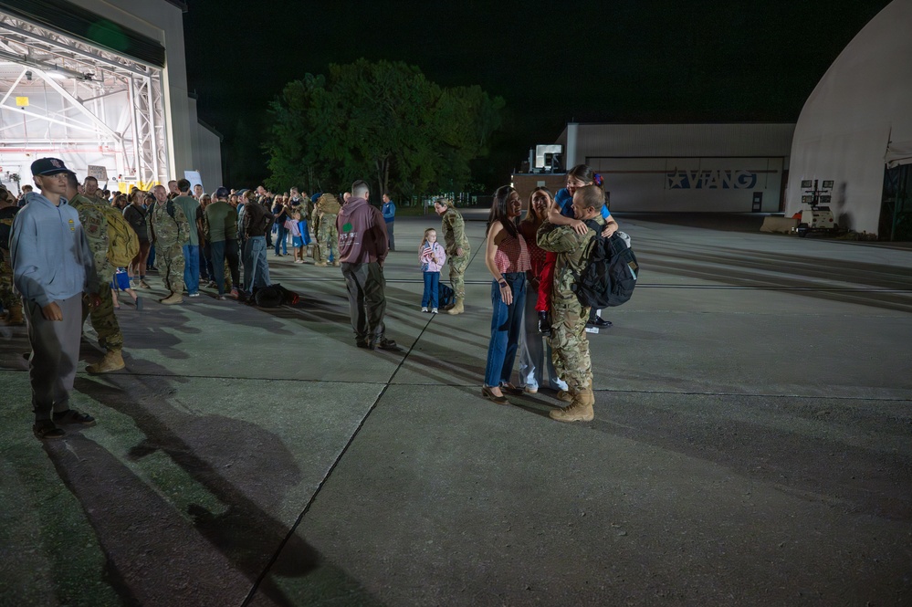 A nighttime military homecoming event on an airfield tarmac, likely at a National Guard base (signage reads 'VIANG' — Virginia Air National Guard). Soldiers in camouflage uniforms are reuniting with family members, including children, near a large illuminated hangar. The scene captures emotional greetings and embraces between returning service members and their loved ones.