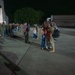 A nighttime military homecoming event on an airfield tarmac, likely at a National Guard base (signage reads 'VIANG' — Virginia Air National Guard). Soldiers in camouflage uniforms are reuniting with family members, including children, near a large illuminated hangar. The scene captures emotional greetings and embraces between returning service members and their loved ones.