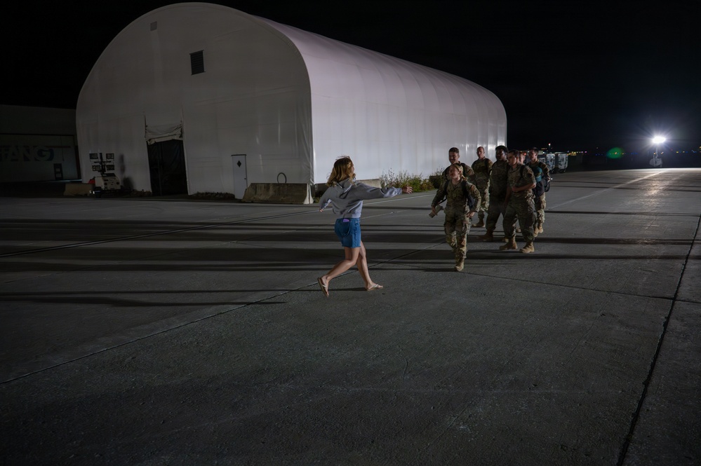 A nighttime scene on what appears to be a military airfield tarmac, where a civilian woman in casual clothing (gray hoodie and denim shorts) is running with arms outstretched toward a group of military personnel in combat uniforms. The setting includes a large white quonset-style hangar structure illuminated in the background. The scene strongly suggests a homecoming or reunion moment.