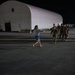 A nighttime scene on what appears to be a military airfield tarmac, where a civilian woman in casual clothing (gray hoodie and denim shorts) is running with arms outstretched toward a group of military personnel in combat uniforms. The setting includes a large white quonset-style hangar structure illuminated in the background. The scene strongly suggests a homecoming or reunion moment.