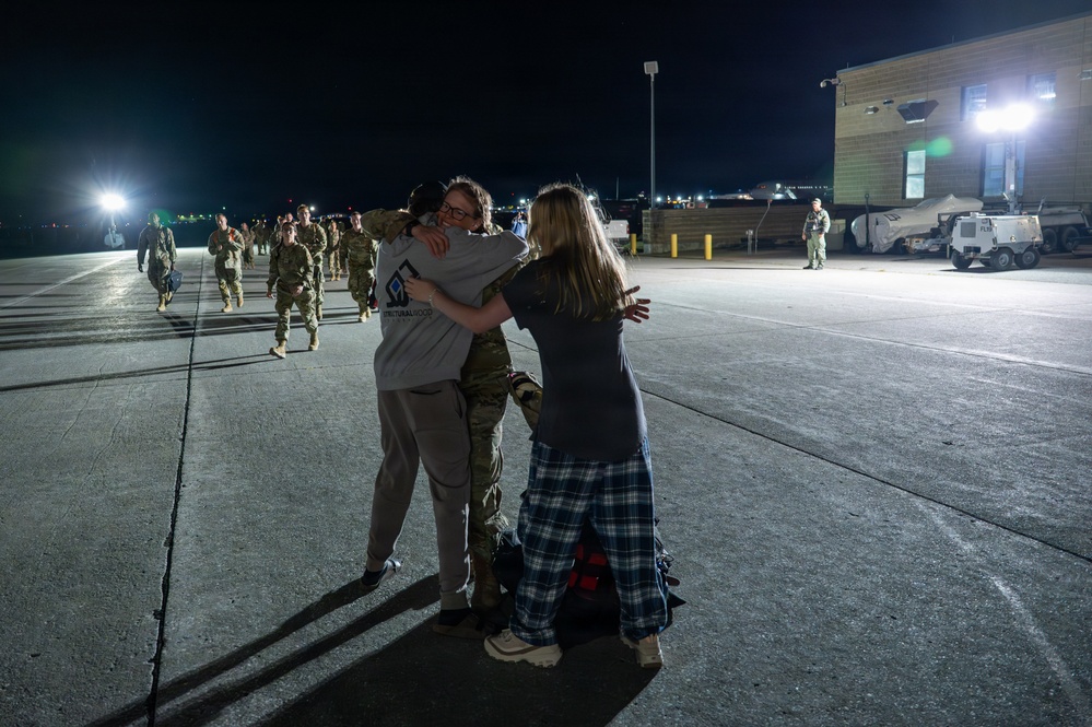 A nighttime military homecoming scene on a tarmac or airfield, where a service member in camouflage uniform is being embraced by two civilians, likely family members. Multiple uniformed military personnel are visible walking in the background under artificial lighting. The emotional reunion appears to be taking place at a military installation.