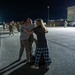 A nighttime military homecoming scene on a tarmac or airfield, where a service member in camouflage uniform is being embraced by two civilians, likely family members. Multiple uniformed military personnel are visible walking in the background under artificial lighting. The emotional reunion appears to be taking place at a military installation.