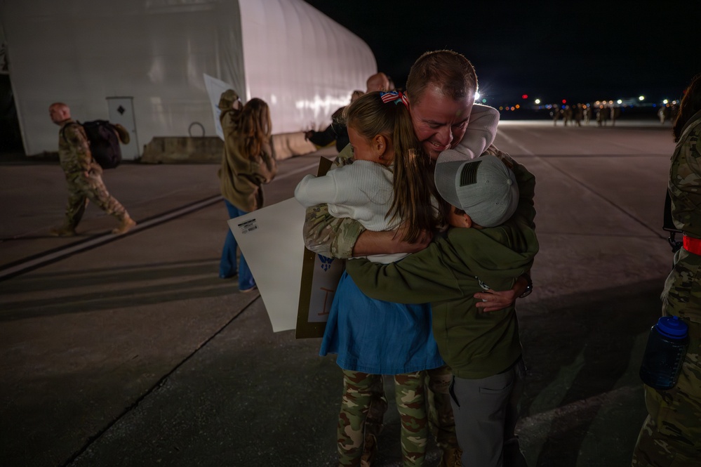 A military service member is reuniting with what appears to be family members, including two children, on an airfield tarmac at night. The emotional embrace shows visible tears on the service member's face, suggesting a homecoming after a deployment. Other uniformed personnel and civilians are visible in the background near a large hangar structure.